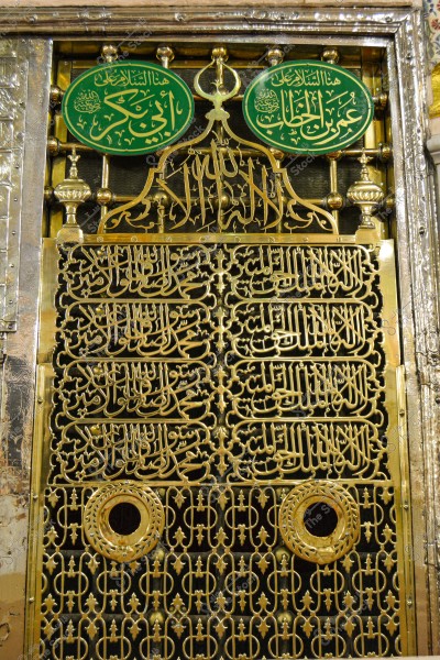Image of an ornate gate made of golden metal, adorned with Arabic inscriptions in calligraphy. The design includes religious inscriptions and intricate elements, representing a significant religious site. Two decorative circles are part of the design, highlighting distinctive architectural features.