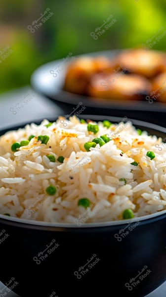An image of a bowl filled with cooked white rice mixed with green peas, placed in a dark-colored bowl. The dish is positioned in the front part of the image, while another bowl appears blurry in the background. The background colors are green and out of focus, suggesting a natural or outdoor setting.