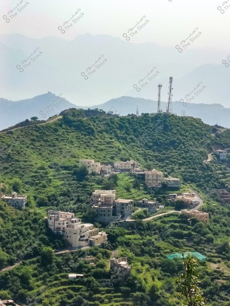 An image of a lush green mountainous area with a cluster of buildings spread across the hills. In the background, a range of mountains adds to the stunning landscape. Some communication towers are concentrated on top of one of the peaks, indicating telecommunications activity in the area. The vegetation is dense, with scattered trees throughout the image.