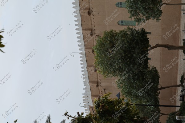 A traditional building with mud walls and green windows, topped with white decorative features on the roof. Green trees are spread in the foreground, and part of the sky is visible in the background. The architectural design suggests a traditional Arab style.