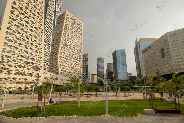 A collection of modern buildings with innovative architectural designs in an urban area. The buildings feature shiny glass and distinctive geometric patterns. The foreground has green spaces and scattered trees, adding natural beauty to the scene. The sky is partly cloudy with soft sunlight reflecting on the glass facades of the buildings.