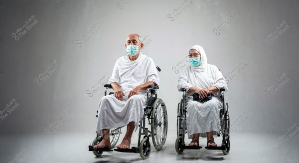 Two elderly individuals sitting in wheelchairs. The person on the left is wearing a white Ihram, typical for Hajj and Umrah rituals, and the other is dressed in a white abaya and headscarf. Both are wearing blue medical masks. The background is grey, highlighting the individuals clearly in the scene.