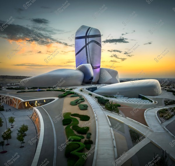 Image of the Ithra building in Saudi Arabia. The building features a unique, futuristic design resembling massive intertwined stones. It is surrounded by manicured gardens and green spaces. The sky in the background is filled with beautiful sunset colors and scattered clouds.