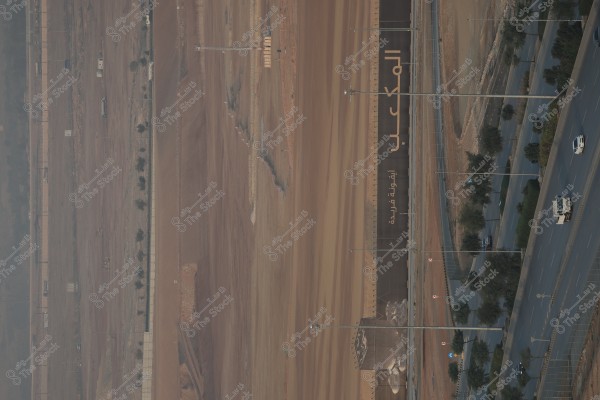 An aerial view of a desert area featuring a large wall with the words "المكعب" and "A unique icon" written on it. To the right, there is a divided road with several cars and trees planted along the edges.