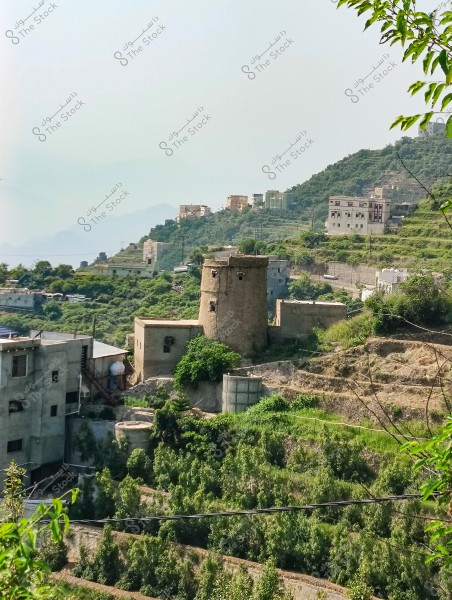 A landscape view of a mountainous area with traditional buildings and terraced agricultural fields in southwestern Saudi Arabia. The image includes stone-built houses, with green hills and scattered trees in the background.