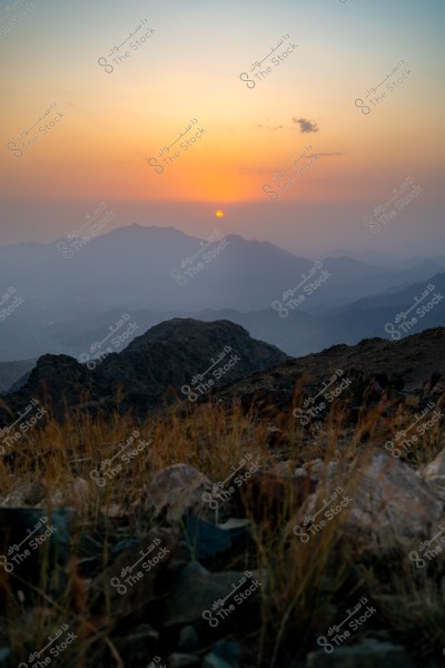 An image of a natural landscape at sunset, with the sun just below the horizon creating an orange sky. Dark mountains stretch across the horizon, and rocky hills covered with tall green plants are in the foreground. The image conveys the tranquility of nature and the beauty of the sunset.