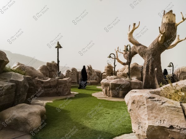 The image shows a natural scene with a grassy path surrounded by large rocks and hollowed trees reaching towards the sky. A woman wearing a black abaya and pulling a rolling suitcase walks along, suggesting she is in an outdoor, possibly tourist, location. Brownish hills form the background, with sunlight illuminating the scene.