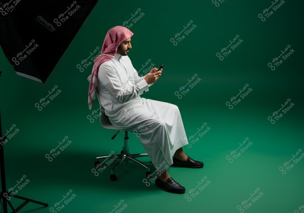 A person wearing traditional Gulf attire, a white thobe, sitting on a chair in a studio setting, viewed from the side. The person wears a red and white checkered headdress and black loafers, holding a mobile phone. A lighting setup is visible in the corner, with a green background.