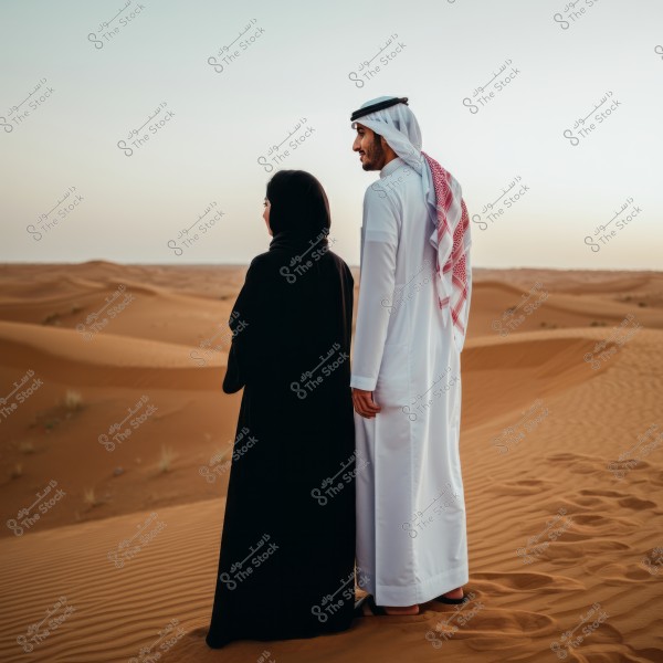 A man and a woman stand in a vast desert, looking towards the horizon. The man is wearing a white thobe and a red ghutra, representing traditional attire, while the woman is dressed in a black abaya and hijab, suggesting a Saudi style. Sand dunes stretch in every direction under a clear sky.