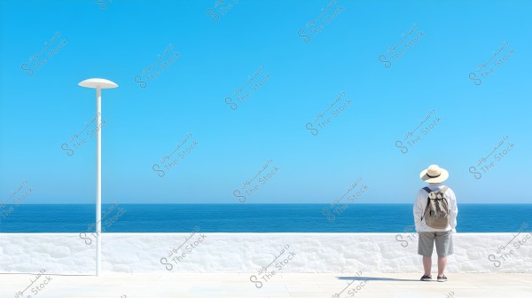 The image shows a person standing in front of the sea, wearing a white shirt, shorts, a wide-brimmed sun hat, and carrying a backpack. Behind them is a low wall and a simple light pole. The sky is clear and blue, with the horizon stretching widely in the background.