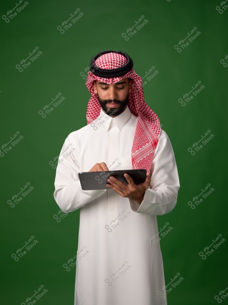 A photo of a man wearing a traditional Saudi white thobe and a red ghutra with a black agal, looking intently at a tablet in his hands. The background is a solid green color.