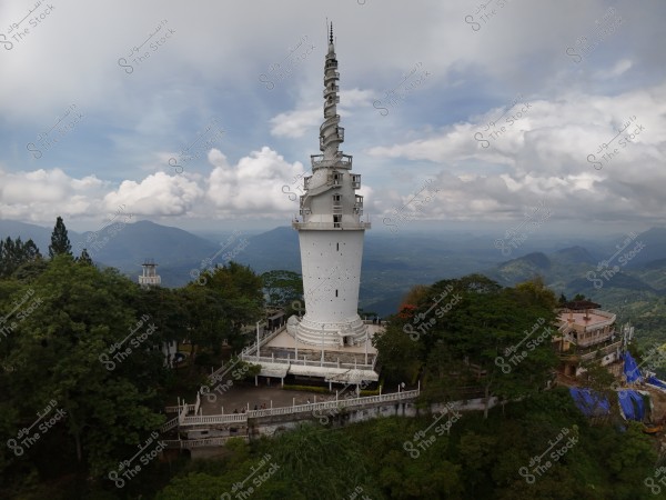 The image shows a tall white tower surrounded by greenery and trees. The tower has a modern design and reaches towards the sky under a blue cloudy sky. In the background, there are mountains and scenic landscapes, providing a serene atmosphere to the location.