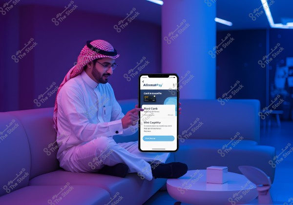 A man wearing traditional Saudi attire is sitting on a sofa in a modern environment lit with blue and purple colors. He is holding a mobile device displaying an app with unclear text. The scene conveys a sense of comfort and modern technology.