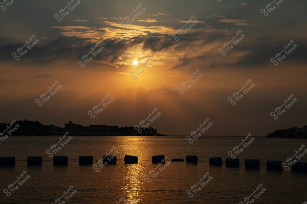 منظر لغروب الشمس فوق البحر, حيث تنعكس أشعة الشمس الذهبية على سطح الماء. السماء مغطاة بغيوم خفيفة تضيف عمقاً وإثارة للمشهد. في الأفق تظهر بعض المباني وأشجار النخيل, وتظهر صورة ظلية مظللة للمباني في مقدمة الصورة.