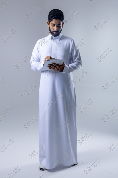 Image of a man standing and wearing a white thobe, resembling traditional Saudi attire, using a tablet with a stylus. The background is white and neutral, giving the image a professional and calm atmosphere.