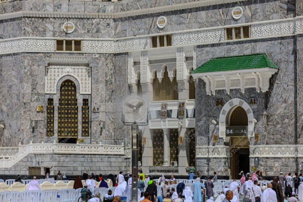 The image shows a section of the Masjid al-Haram in Mecca, Saudi Arabia. The facade features marble with traditional Islamic architectural details and ornate arches. Several people wearing white Ihram clothing and others in various traditional attire are gathering in the surrounding courtyard.