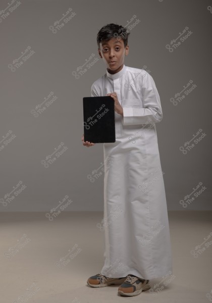 A young boy stands in a studio wearing traditional Saudi white attire, holding a tablet. The boy is dressed in a white thobe and sports shoes, with a simple gray background behind him.
