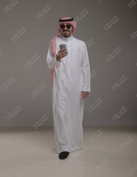 An image of a man wearing a white thobe and a red checkered headdress with an agal. He is wearing sunglasses and holding a mobile phone, standing with a slight smile on a grey background. The attire appears to be traditional Gulf clothing.