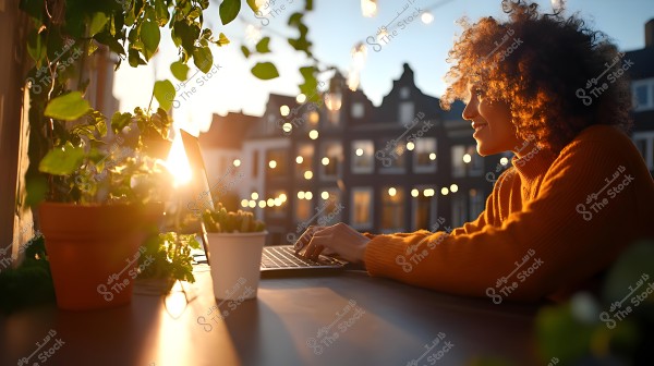 A woman in an orange sweater sits on an open terrace working on a laptop during sunset. Green plants surround the table, and string lights hang in the background, with a residential building and a large window behind her.