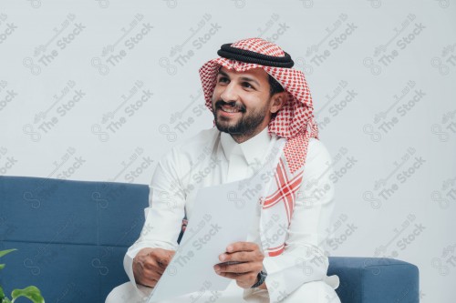 رجل سعودي مبتسم يرتدي الزي السعودي التقليدي وممسك بورقة لمناقشتها أثناء اجتماع عمل، موظف أثناء العمل في غرفة الاستراحة