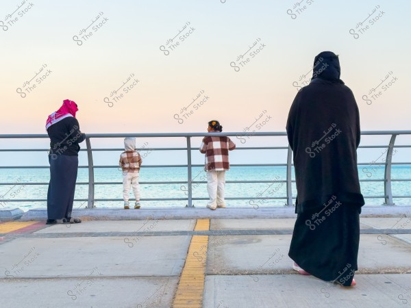 صورة لمشهد عائلي على الواجهة البحرية. يظهر في الصورة رجل يرتدي الغترة والعقال وامرأة ترتدي العباءة السوداء بالإضافة إلى طفلين يرتديان ملابس شتوية واقفين عند السور الحديدي ويطلون على البحر, مع منظر السماء الصافية في الخلفية.\r\n\r\n-