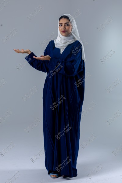 A portrait of a woman wearing a dark blue abaya and a white headscarf, looking at the camera with a slight smile, and holding her hands up in an open gesture. The background is a neutral light gray.