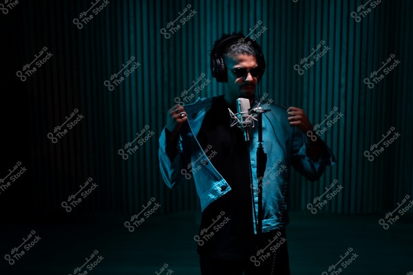 رجل يرتدي نظارات شمسية وسماعات رأس ويغني أمام ميكروفون في استوديو مضاء بشكل خافت.