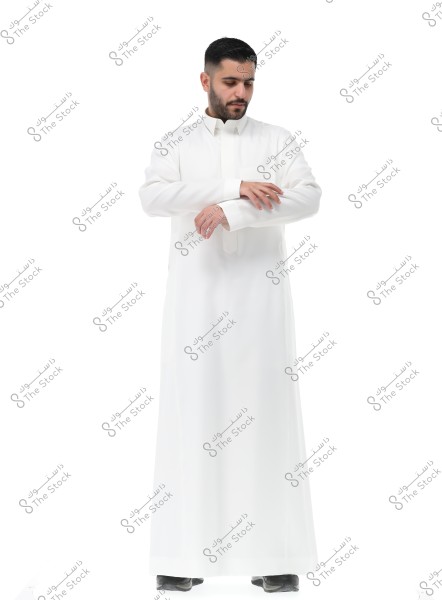 The image shows a man wearing a full white thobe standing against a white background. The man appears to be adjusting the sleeves of his thobe. The thobe represents traditional clothing commonly seen in Gulf countries like Saudi Arabia. The man has a short beard and dark hair.