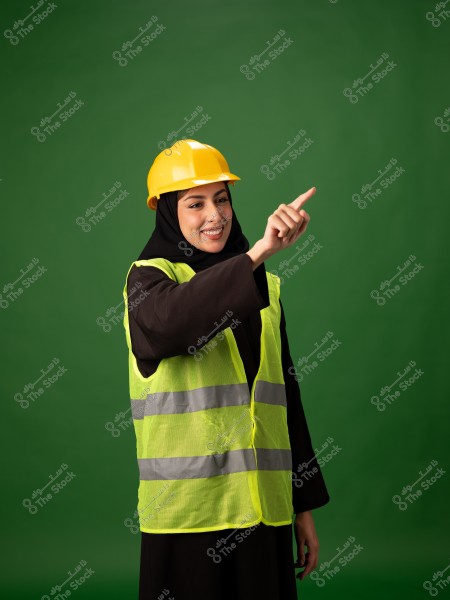 Image of a woman wearing a yellow hard hat and a bright green reflective safety vest with silver stripes, pointing to the right with a smile. She is wearing a black abaya and a black hijab. The background is a solid green.