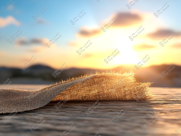 An image showing a piece of burlap placed on a wooden surface, under the glow of the sun on the horizon. The golden sun appears to be setting behind mountains in the background, casting a warm reflection on the fabric and creating beautiful light effects.