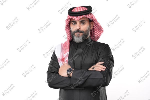 A portrait of a man standing with his arms crossed over his chest. He is wearing a black thobe and a white and red checkered ghutra with an agal. The man has a serious expression and the background is white, suggesting a formal portrait. The attire indicates the man is likely from Saudi Arabia or another Gulf region.