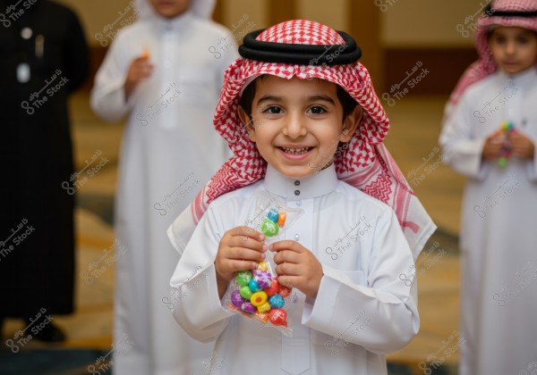 An image of a young boy wearing a white thobe and a red and white checkered ghutra, smiling and holding a transparent bag filled with colorful candies. In the background, other children dressed in similar traditional attire are visible, suggesting a cultural or festive occasion. The image likely originates from Saudi Arabia.