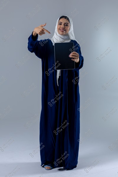 امرأة ترتدي عباءة زرقاء وحجابًا أبيض تقف على خلفية رمادية. تحمل بيد واحدة جهاز لوحي أسود فارغ وتستخدم اليد الأخرى للإشارة إليه.