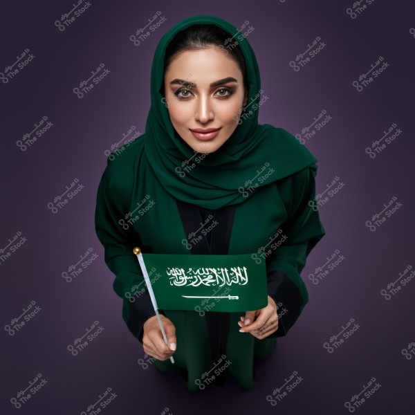 A portrait of a woman wearing a green hijab and abaya, holding a small Saudi Arabian flag in her hand. The background is a dark purple color. The flag displays the shahada and a sword.