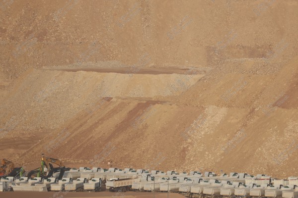 The image shows a construction or mining site in a desert area with rocky terrain. The bottom part of the image features several large trucks lined up alongside a number of excavators, indicating construction or transportation activity at the site.