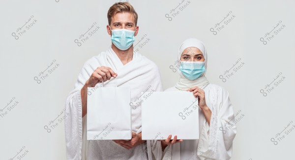 A photo of a man and woman standing side by side, both wearing white Ihram clothing, indicating they are in Saudi Arabia. The man holds a white shopping bag while the woman holds a white sign. Both are wearing blue face masks as a precautionary measure.