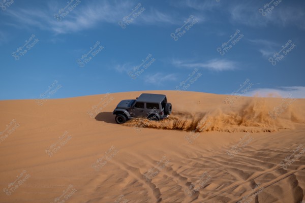 A four-wheel-drive vehicle drives across golden sand dunes in the desert, kicking up clouds of sand behind it. The clear blue sky in the background adds to the beauty of the scene.