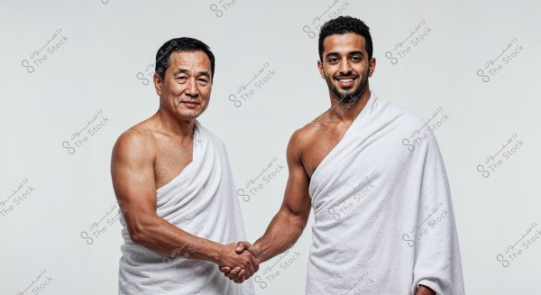 Two men wearing white Ihram garments, shaking hands with smiles. The man on the left has East Asian features, while the man on the right appears to have Middle Eastern features. They are standing against a white background.