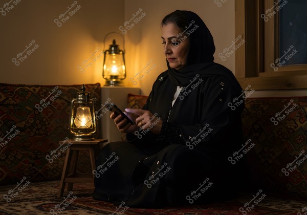 A photo of a woman sitting in a room illuminated by warm yellow lights from traditional lanterns. She is wearing a black abaya and hijab, using a smartphone. The background features traditional Arabic patterns and a wooden corner of the room.