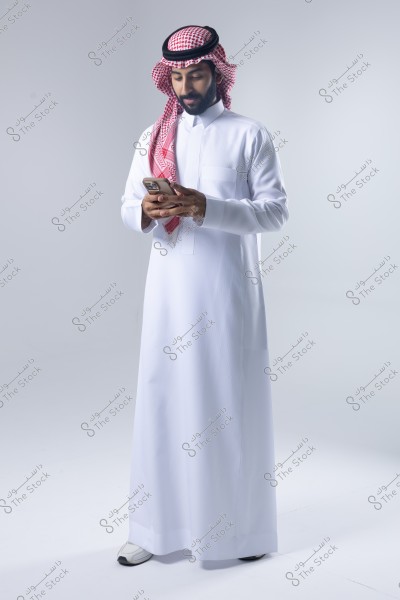 صورة لرجل يرتدي ثوبًا أبيض وغترة حمراء مع عقال أسود, ويستخدم هاتفًا ذكيًا. الخلفية عبارة عن مساحة رمادية بسيطة دون تفاصيل إضافية. يبدو أن الرجل من أصل خليجي حيث أن الملابس تقليدية في دول الخليج.