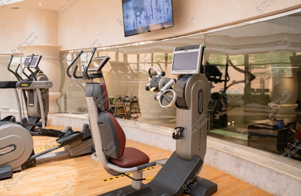 The image shows the interior of a modern gym featuring advanced workout machines. In the foreground, there is an exercise machine with a large display screen and circular handles, with a background of reflective mirrors on the wall. Additional exercise equipment is visible in the background, and the floor is made of wood.