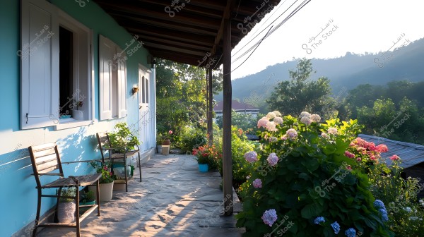 An image of a house porch adjacent to a blooming garden. The walls are painted light blue with open white windows and wooden chairs arranged on a stone pavement. The porch overlooks a natural landscape filled with trees, various flowers, and mountains in the background under sunlight.