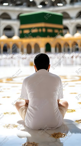 A person sitting on the ground in the courtyard of the Grand Mosque in Mecca, wearing white Ihram clothing. In the background, the Kaaba is visible, surrounded by decorated marble walls.