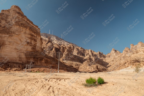 An image of a mountainous area with towering sandstone cliffs under a clear blue sky. In the middle of the image, there are some electric poles and power lines, while some green shrubs are growing in the sandy desert foreground.