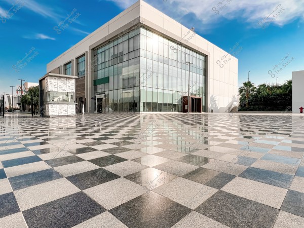 A modern building with a large glass facade located in the middle of a wide plaza covered with black and white checkered tiles. The sky is clear and blue, with some trees in the background.