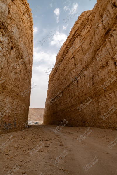 An image of a narrow sandy path between two high cliffs of sandstone, with a blue sky and some clouds in the background. The image reflects a desert environment and rocky geography.