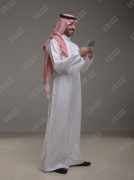 A portrait of a man wearing traditional Saudi attire, featuring a white thobe, a red checkered headscarf, and a black agal. The man is smiling and looking at his smartphone, which he holds with both hands. The background is a plain gray, suggesting a studio setting.