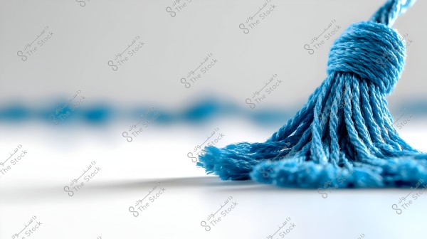 A close-up image of a blue tassel made of threads placed on a white surface with a blurred background. The tassel is tightly wrapped and knotted at the top, with the lower threads spreading loosely.
