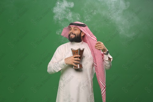 رجل سعودي يقف على خلفية خضراء يرتدي الثوب و الشماغ، يقوم بعمل حركات بيديه ، يبخر نفسه بالطيب و العود، احتفالات العيد