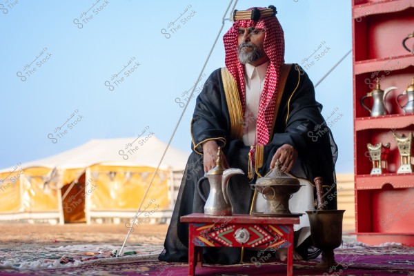 An image of a man sitting in a traditional tent, wearing traditional Saudi attire, including a red and white headscarf and a black decorated bisht. In front of him is a small decorative table with traditional coffee-making tools. In the background is a white tent and several traditional coffee pots on a red shelf.